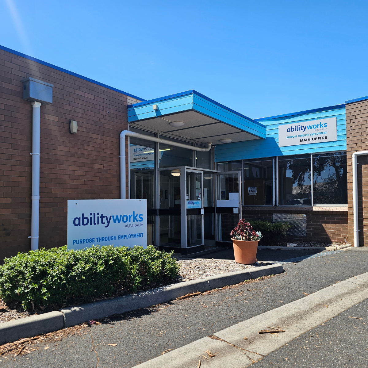 External view of the Ability Works office entrance. There are signs with the company logo, the building is made of brick and there is a section of blue roofing above the entry.